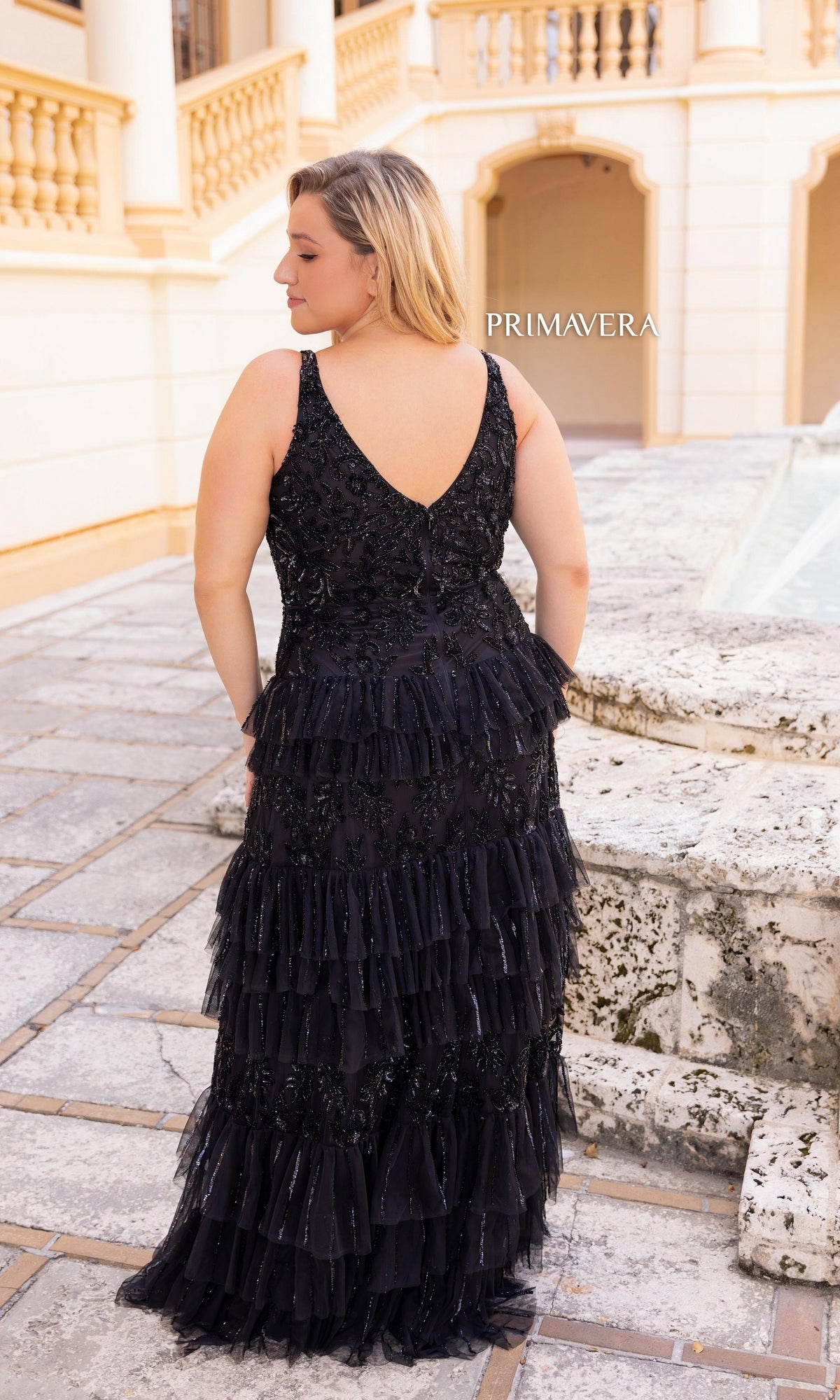 A woman in the Primavera Tiered Plus-Size Long Prom Dress 14052, featuring a sleeveless black beaded design with tiered ruffles, stands outdoors near a stone fountain, facing away from the camera.